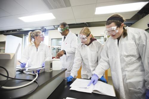 Chemistry students, wearing goggles and lab gear, work with a professor in a chemistry lab.