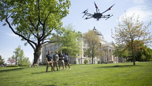 A drone flies near Academic Hall as a student pilot and peers stand nearby.