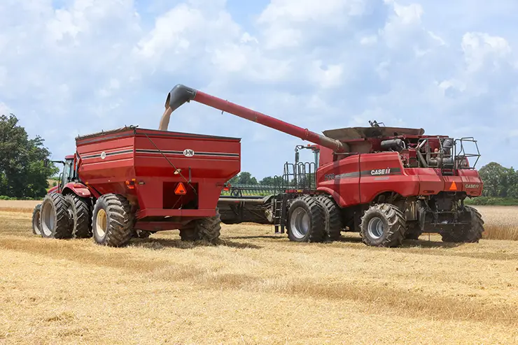 a harvester machine and a tractor with a wagon sit side by side in a wheat filed while the harvester unloads