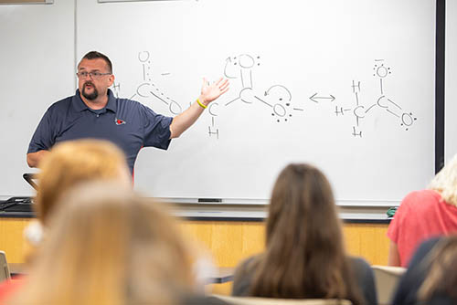a chemistry instructor stands at the head of the class and describes a formula