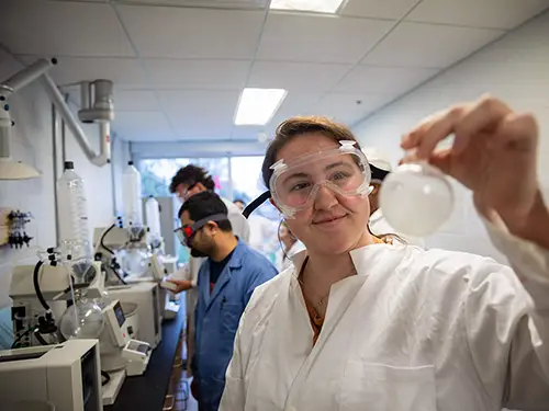 a chemistry and math student holds up a piece of lab glass with a row of equiment behind her