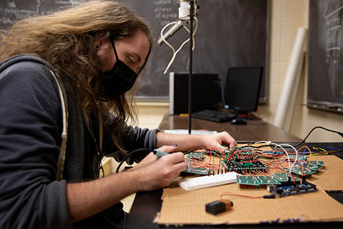 a student is seated at a desk with circuit boards and wires spread out in front of him on top of a piece of cardboard