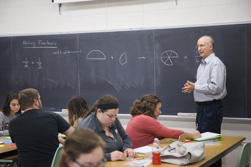 Students take notes while a professor explains the concept of adding fractions and pie charts on the chalkboard.  