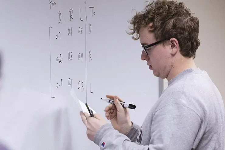 a math student stands at the white board with a paper in his hand writing an equation