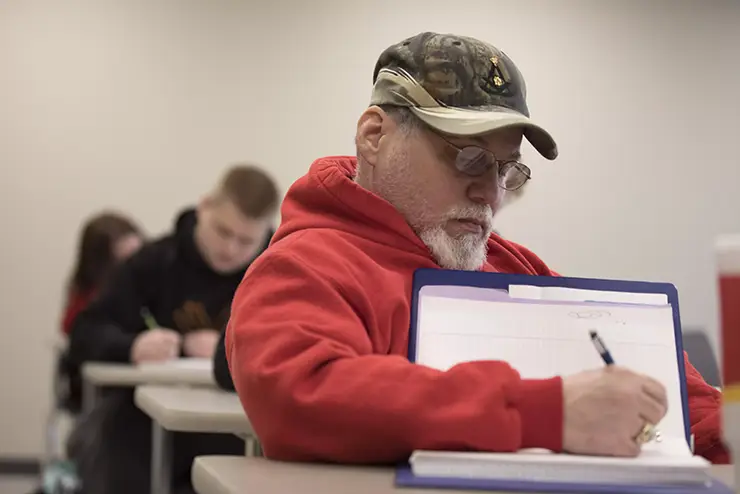 an older, non-traditional student sits in a classroom taking notes on a pad of paper