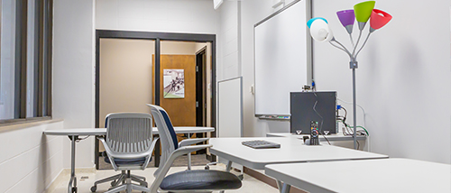Classroom with windows and a white board on the wall. Desks and chairs scatter the small room.