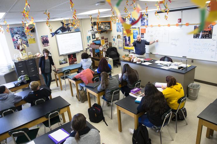 A young man in a blue shirt points at a whiteboard while a group of students look on.