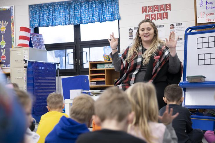 A young woman wearing a black and red checkered scarf holds her hands up as she directs a small group of children.