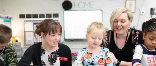 A  young woman wearing a white dress with a black sweater works with a young child while a mentor teacher looks on.