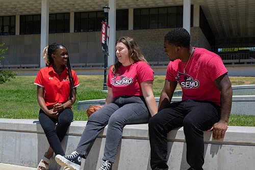 Three students dressed in Redhawk gear conversing in front of Kent Library. 