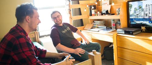 Roommates laugh and hangout to play a video game on an Xbox in their dorm room as the sun shines brightly through their window. 