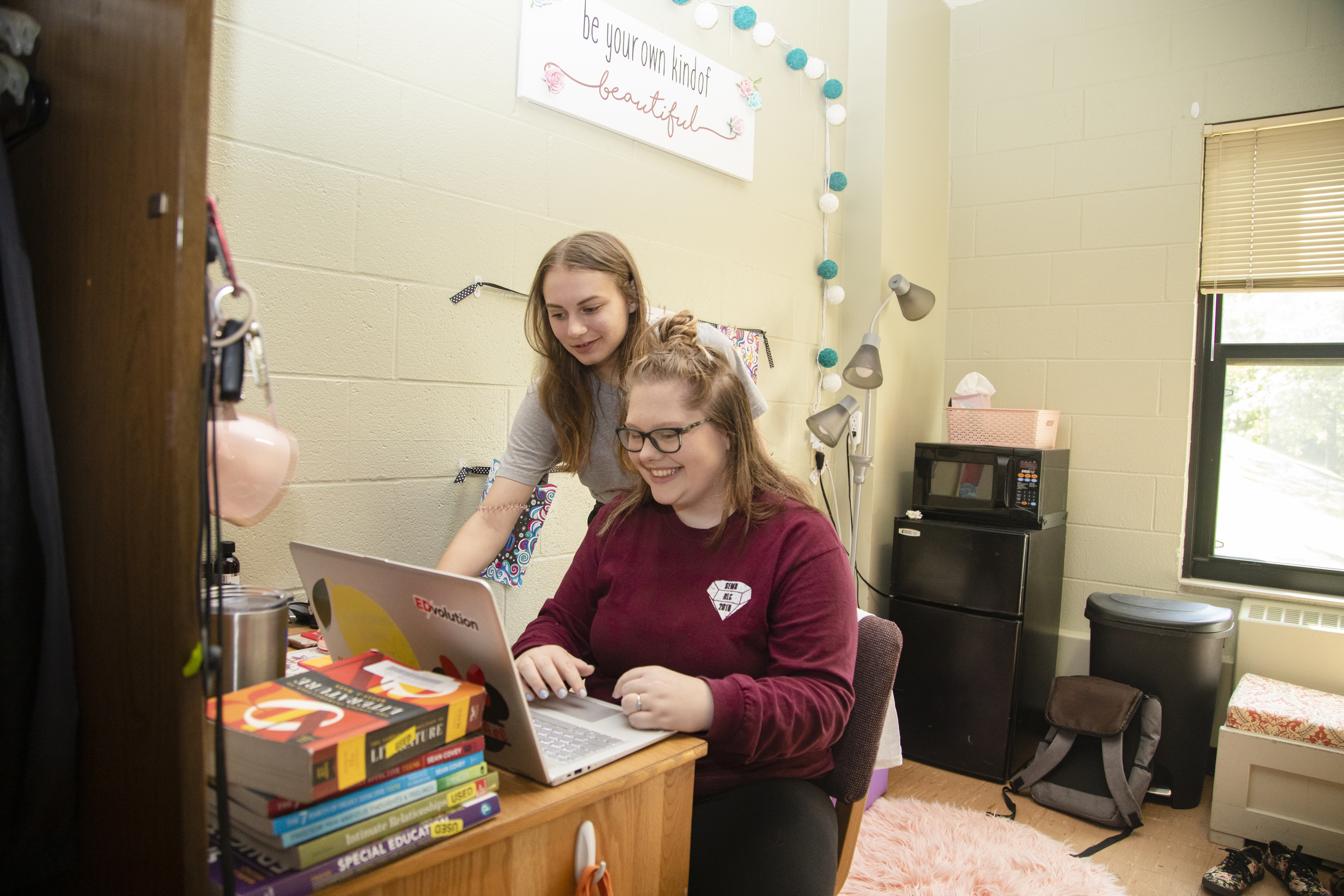 Two semo students who are roommates look at a laptop on a desk in their dorm on campus. The space is decorated and includes a desk and MicroFridge. 