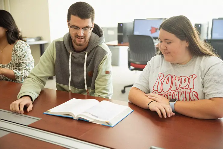 two SEMO students sit together at a table with a book open between them