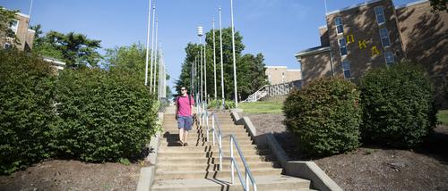 A student in cargo shorts, a red striped polo, and sunglasses walks down the steps of Greek Hill with his hand in his pocket by residence halls with Greek letters on them.  