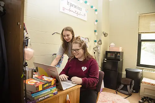 Two SEMO students who are roommates look at a laptop on a desk in their dorm on campus. The space is decorated and includes a desk and MicroFridge. 