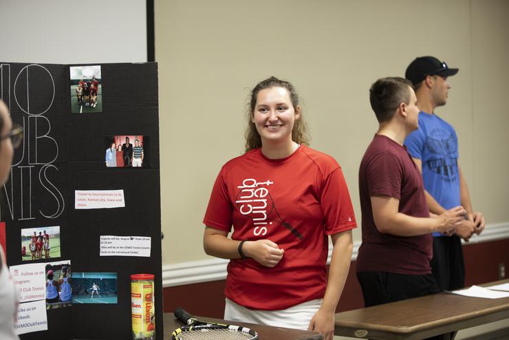 A student in a tennis club t-shirt waits to talk with involvement fair attendees. 