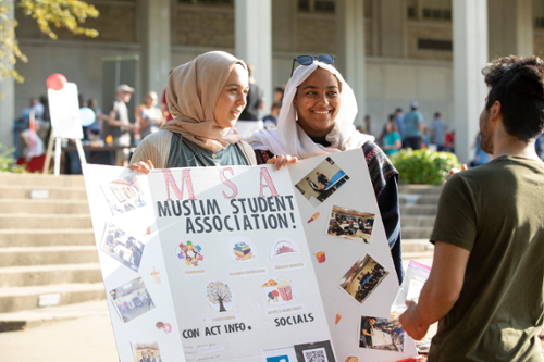 Two students are holding a poster for Muslim Students Association while talking to another student.