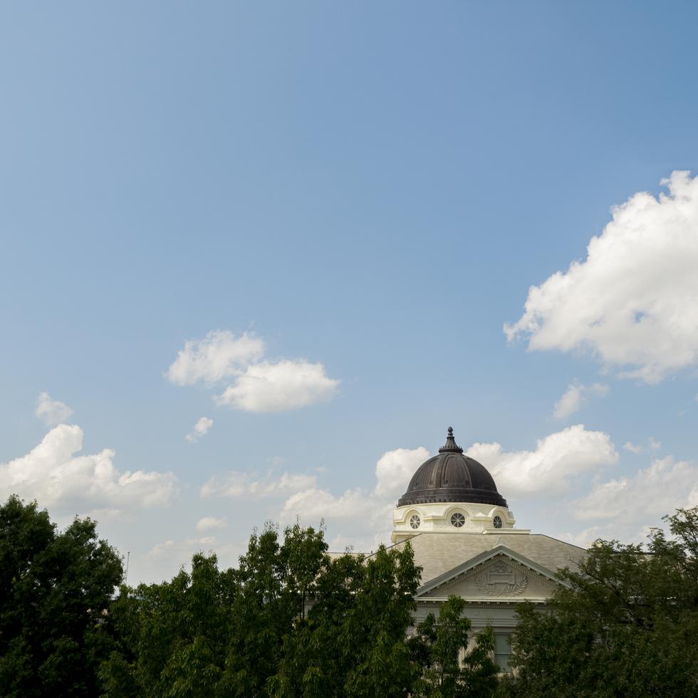 The dome of Academic Hall shows above the tree line on a bright day with clouds in the sky.  