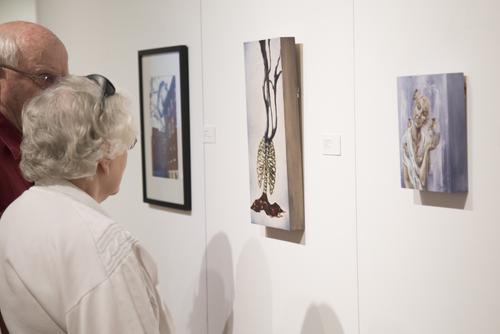 Two older individuals stand with their backs to the viewer, looking at canvases hung on the gallery wall.