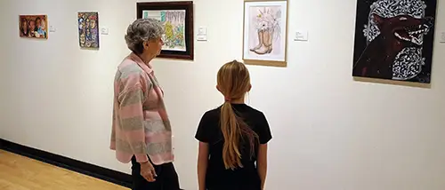 two patrons of Crisp Museum stand in the gallery and view paintings on display