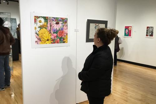 a museum patron in a dark jacket is standing at the gallery wall looking at a piece featuring colorful flowers