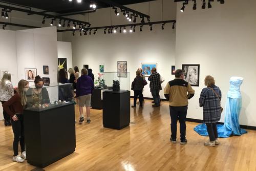 a wide view of a part of Crisp Museum full of visitors with artwork on stands under glass, in walls and a dress on a mannequin
