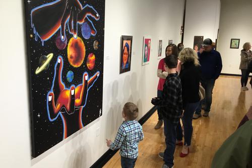 groups of museum visitors look at artwork on the walls, in the foreground is a colorful painting of hands cupping a world