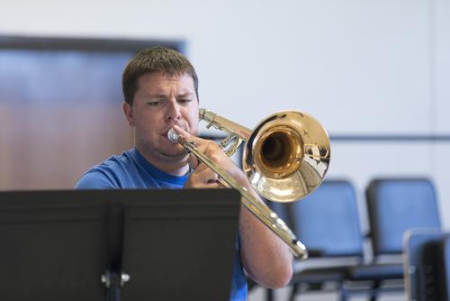 A Music Academy member plays a trombone while looking at a sheet of music on a stand.