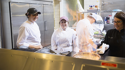 Students watch in a kitchen as an instructor sautees a dish that flames up. 