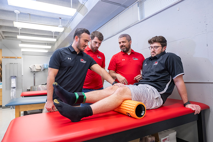 SEMO students learning athletic training from Dr. Kyle Schneider in a white brick room on a red seat.