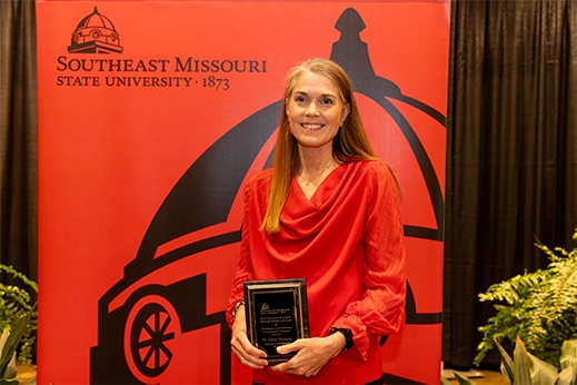 A faculty member is presented with a black plaque award in front of a red banner.