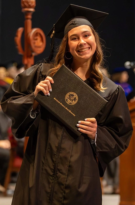 A young woman in her regalia smiles with her diploma at commencement.