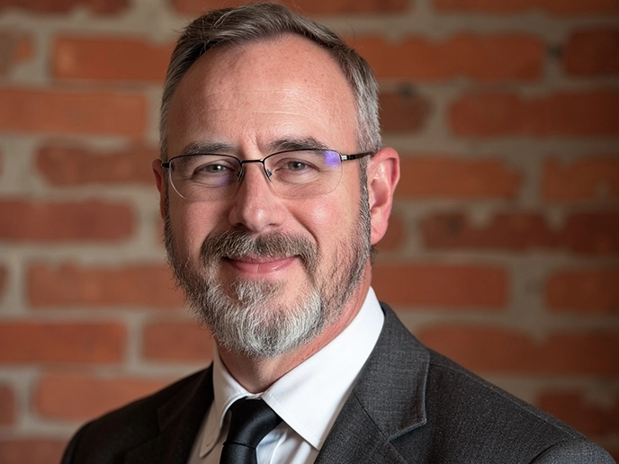 A professional headshot of a man in a dark suit in front of a brick background.