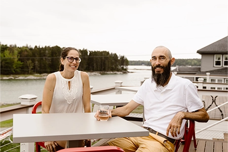 A man and woman sit at a high top table on a balcony overseeing a body of water.