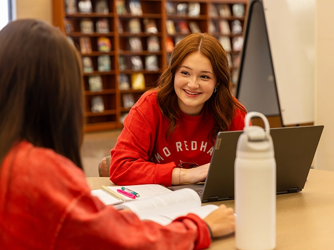 A student talks with her friend at a desk.