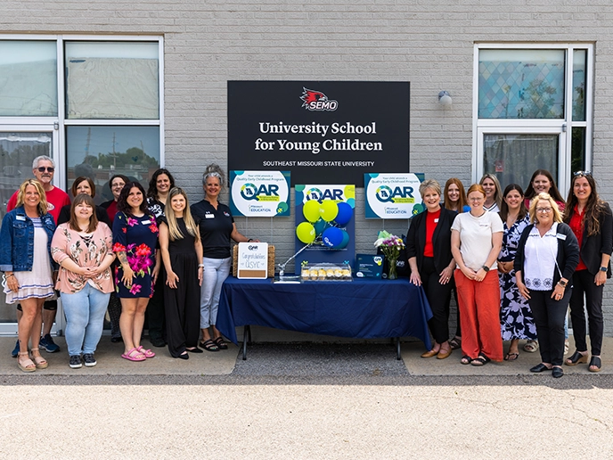 A group stands beside a table with blue and green signage outside a gray building with a black sign.