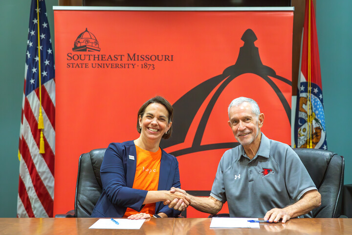 Wyman CEO Allison Williams shaking hands with SEMO President Dr. Carlos Vargas in front of a red SEMO banner and the American and Missouri state flag.