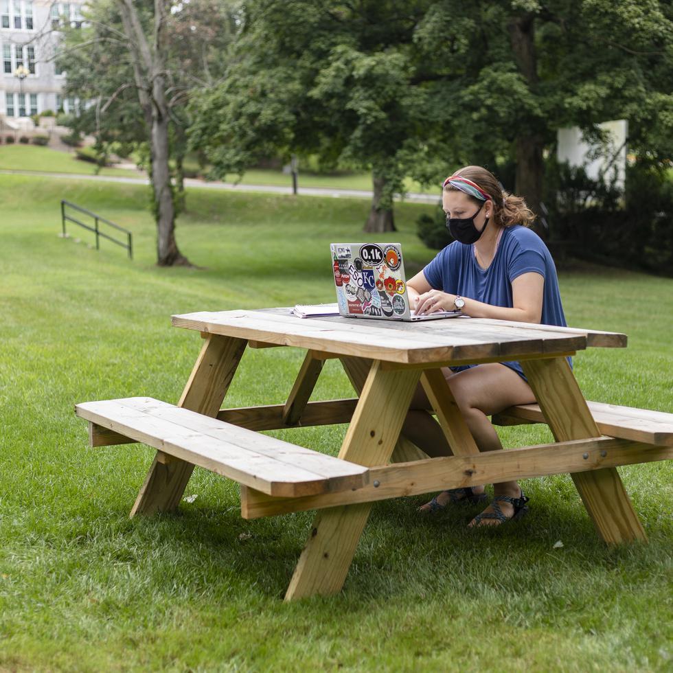 A student works on their laptop at a wood picnic table in the lawn area near the Academic Terraces.  