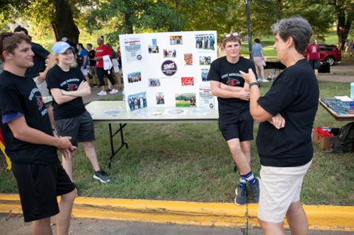 Three student members of Sports Management Society talk with a faculty member while standing around a poster board.