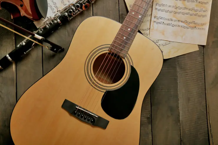 A guitar next to a flute, violin, and sheets of music on wood flooring.