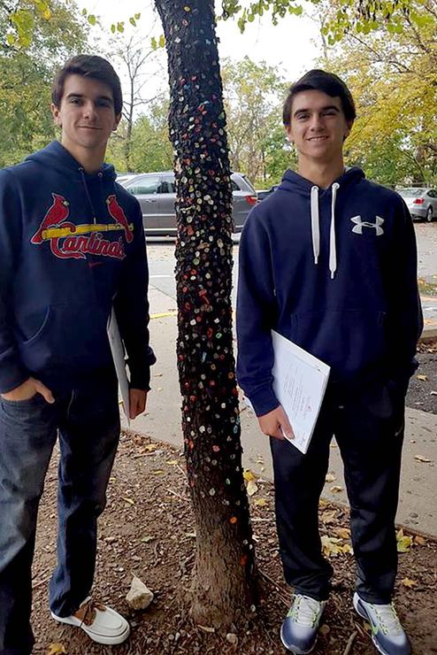 Brandon and Collin Roth, both wearing navy sweatshirts, pose by the Gum Tree, its trunk covered in colorful gum.