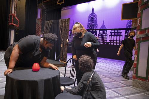 Students gathered around a table set piece, getting into place for rehearsal