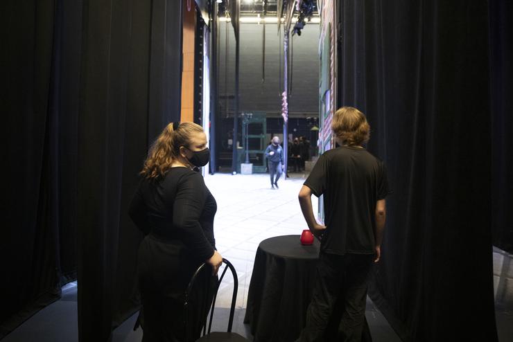 Two students wearing all black stand in the wings, ready to bring props on stage.