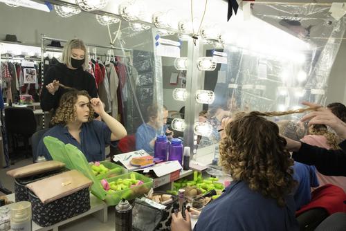 A member of the wardrobe crew curls an actresses hair as the actress does her own make up.