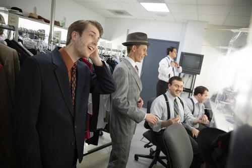 group of male students in the dressing room backstage talking and laughing and applying makeup