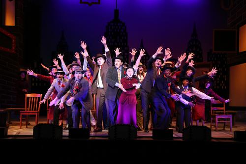 cast of Guys and Dolls grouped all together on stage with their hands in the air performing a song and dance piece