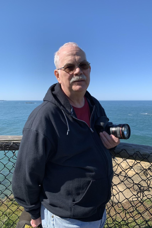 Ron stands with his camera near a body of water.