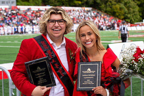 A man and woman stand and smile together, holding flowers and awards