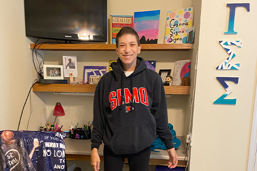 A girl in a SEMO sweatshirt smiles in her dorm room