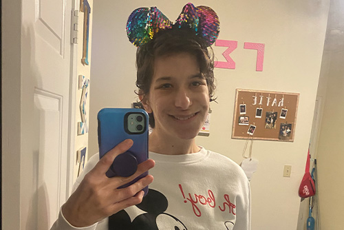 A girl takes a selfie with her phone while wearing Mickey Mouse ears in the mirror
